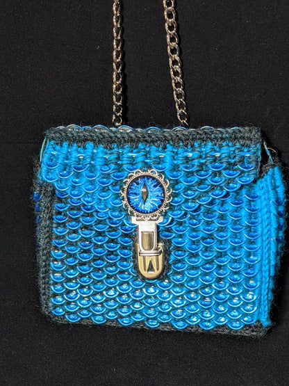 Small dragon scale bag hand crocheted using upcycled can tabs and a wool blend yarn featuring  a hand painted glass dragon eye on the flap. Bag pictured is blue and dark grey.